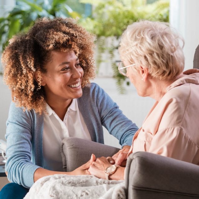 Female home caregiver talking with senior woman, sitting in living room and listening to her carefully.