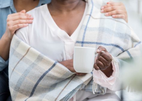 Unrecognizable woman comforts her friend who is ill. She gives her a warm blanket and a cup of hot tea.
