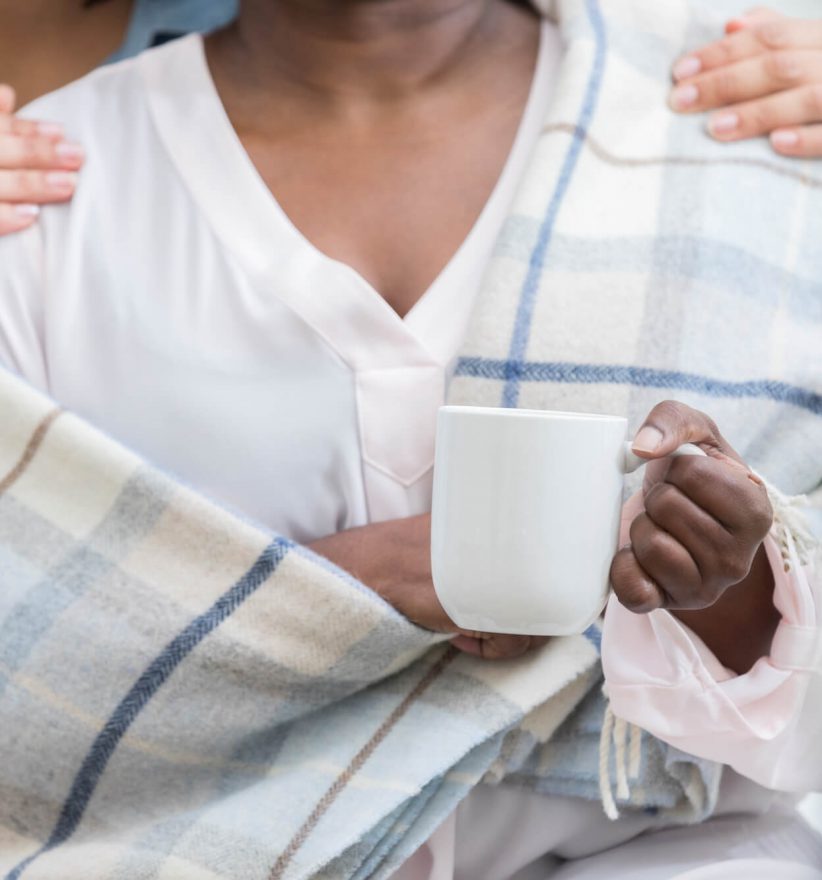Unrecognizable woman comforts her friend who is ill. She gives her a warm blanket and a cup of hot tea.